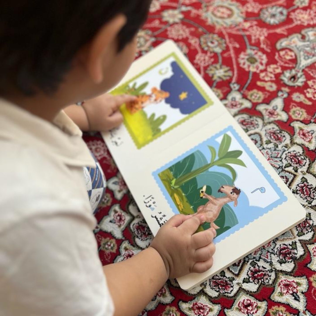 A toddler sitting on a patterned rug reading an Arabic board book, pointing at illustrated animals and Arabic letters on colorful pages.