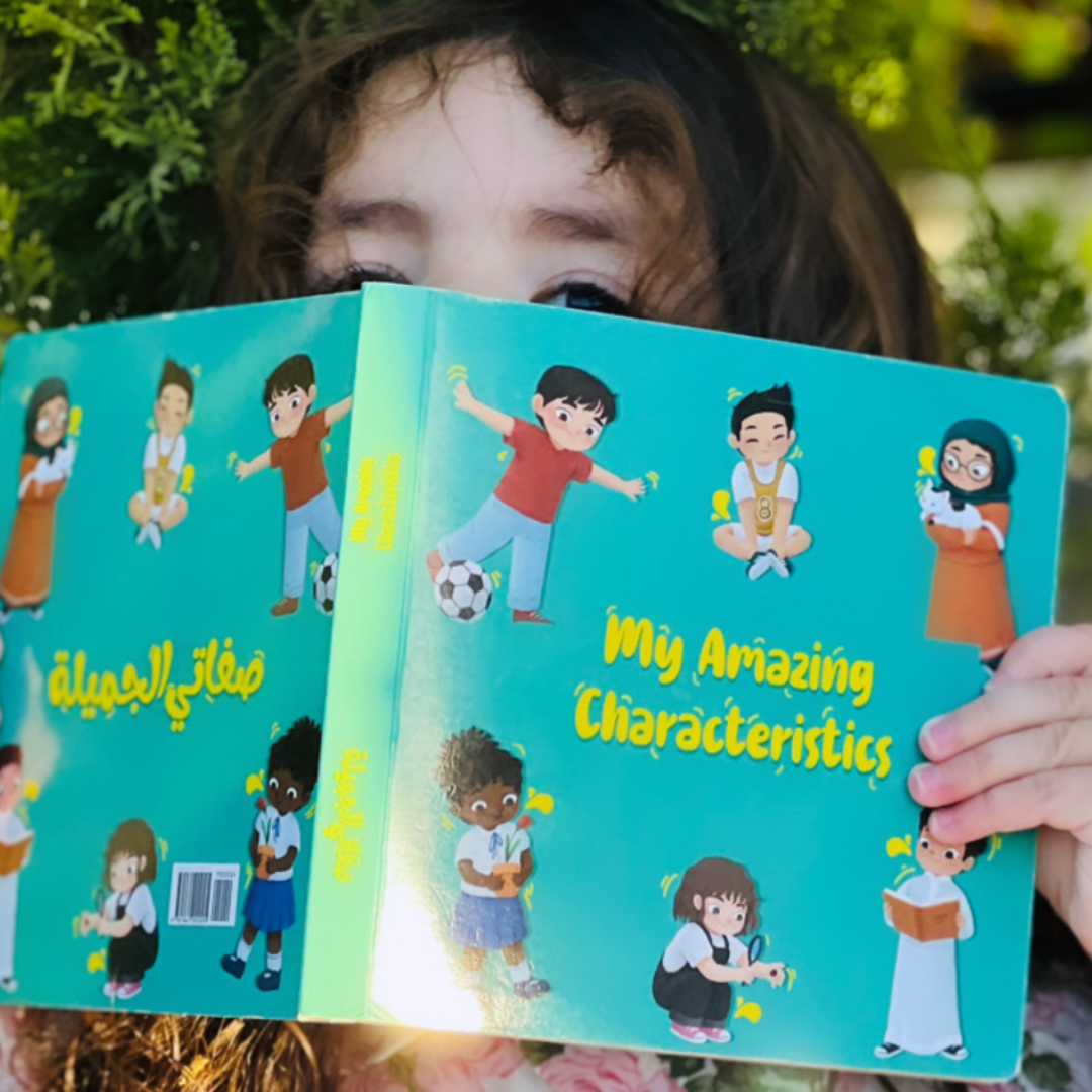 A young child holds up the bilingual board book “My Amazing Characteristics” outdoors, showing its colorful Arabic-English cover featuring diverse children.