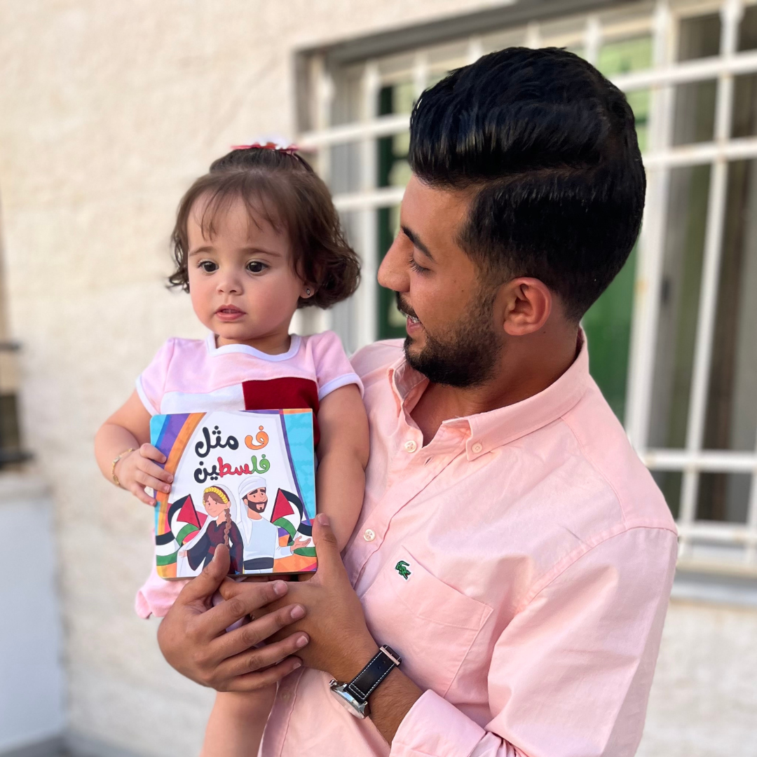 Father holding his toddler daughter as she holds the Palestinian Arabic Alphabet Book, “ف مثل فلسطين,” outside their home.