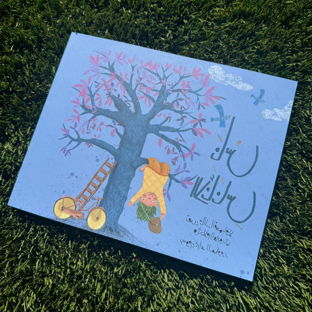 Photo of “The Pineapple People” Arabic children’s book lying on grass. The cover features a child with green hair hanging upside down from a tree with pink leaves, next to a yellow bicycle and ladder, with Arabic title text.
